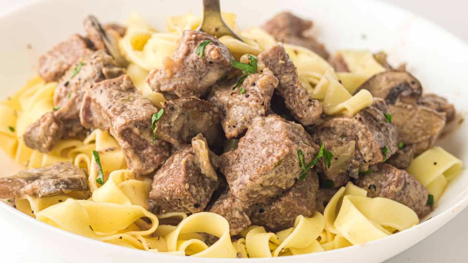 A bowl of egg noodles topped with creamy beef and mushroom stroganoff, garnished with chopped herbs.