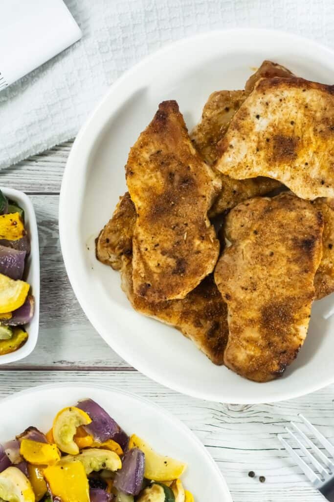Broiled Pork Chops on a white plate, seasoned and golden brown. Served with colorful roasted vegetables, creating a hearty, rustic meal setting.