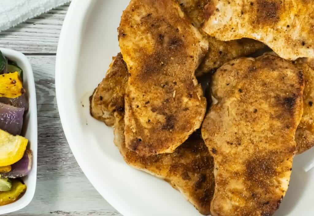 Broiled Pork Chops with a golden brown crust on a white plate, accompanied by a dish of colorful roasted vegetables in the foreground.