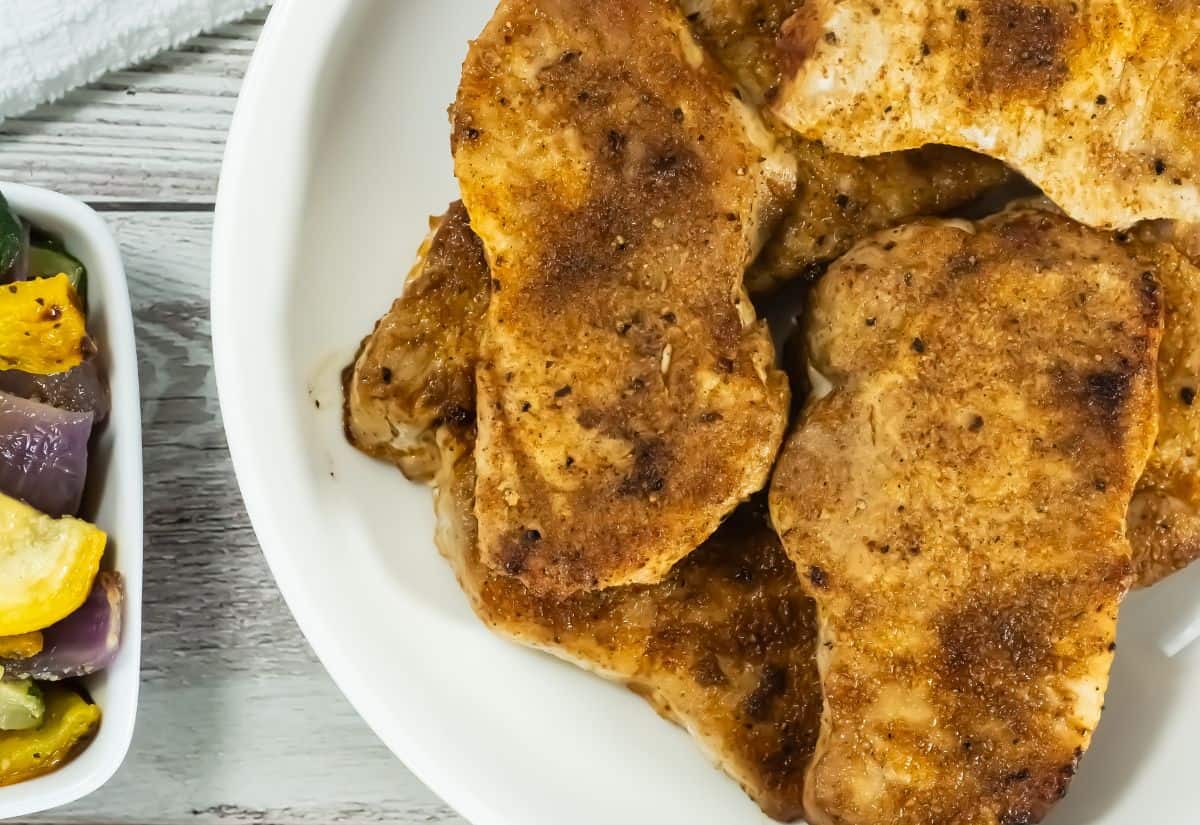 Broiled Pork Chops with a golden brown crust on a white plate, accompanied by a dish of colorful roasted vegetables in the foreground.