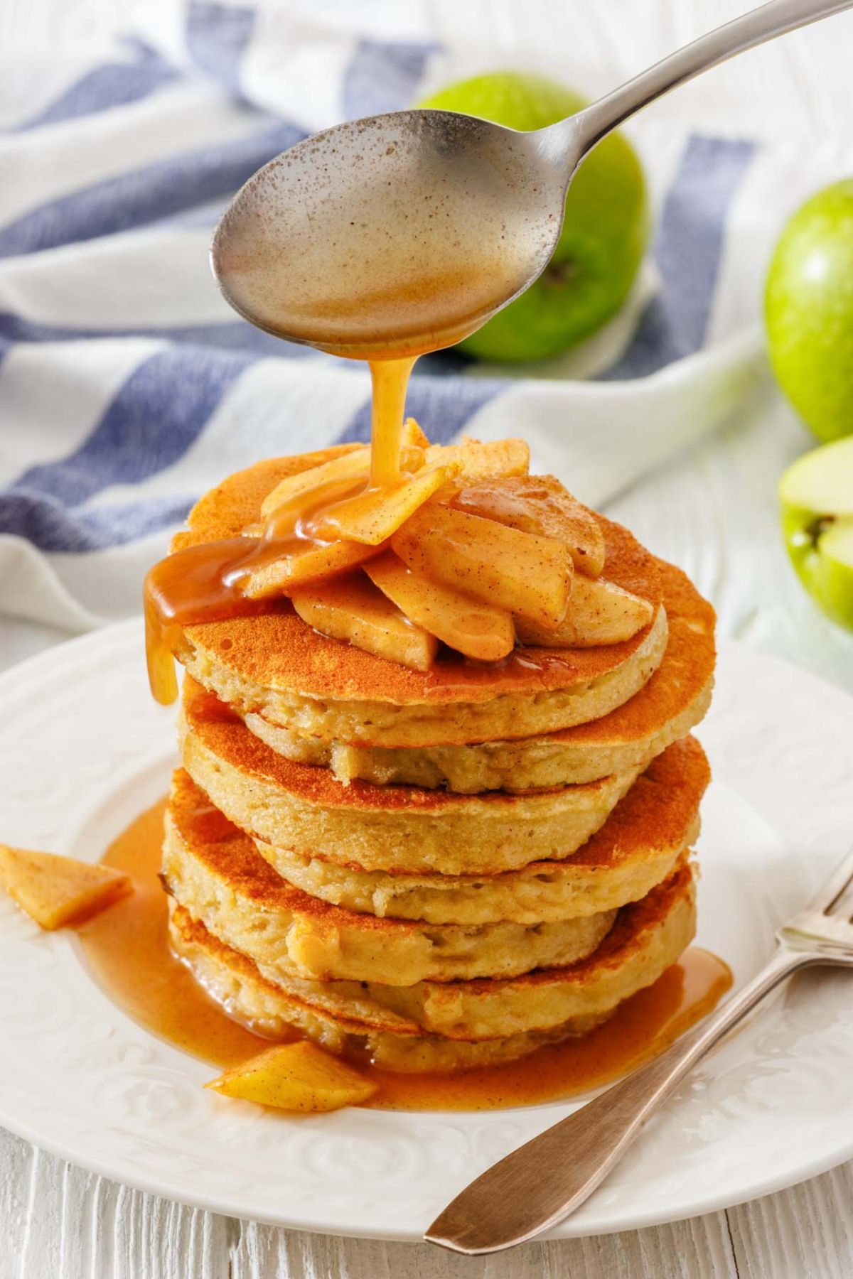 A stack of pancakes topped with caramelized apple slices is being drizzled with syrup from a spoon, set on a white plate with a striped cloth nearby.
