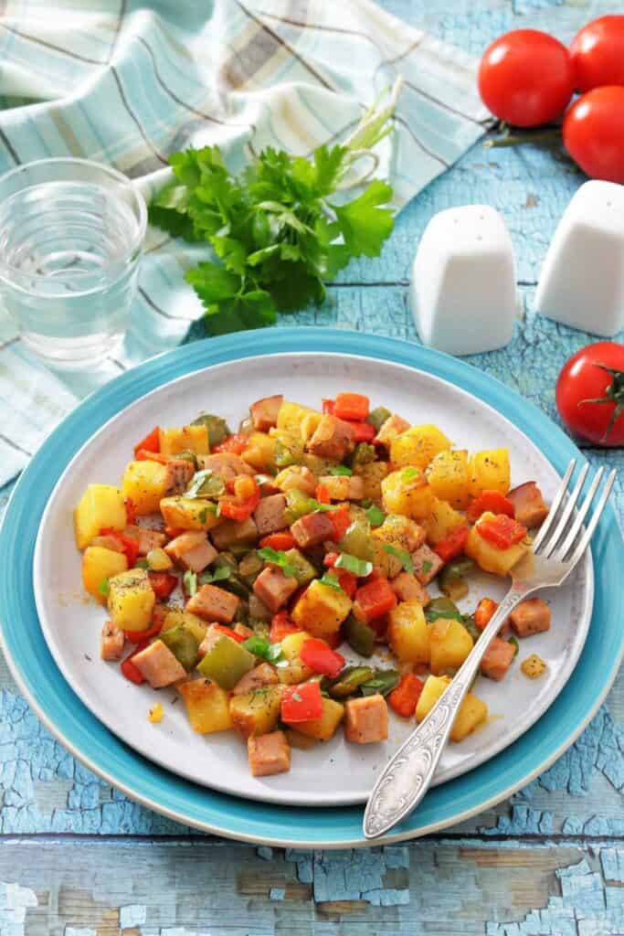 A vibrant, colorful vegetable and meat hash served on a white plate with a blue rim. Surrounded by cloth, tomatoes, parsley, and a glass of water.