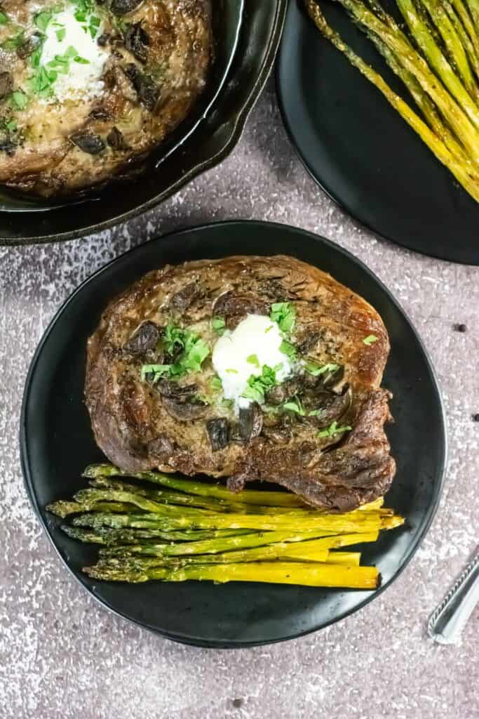 Grilled steak topped with butter and herbs on a black plate, paired with roasted asparagus. Another dish with asparagus is partially visible. Rustic tone.