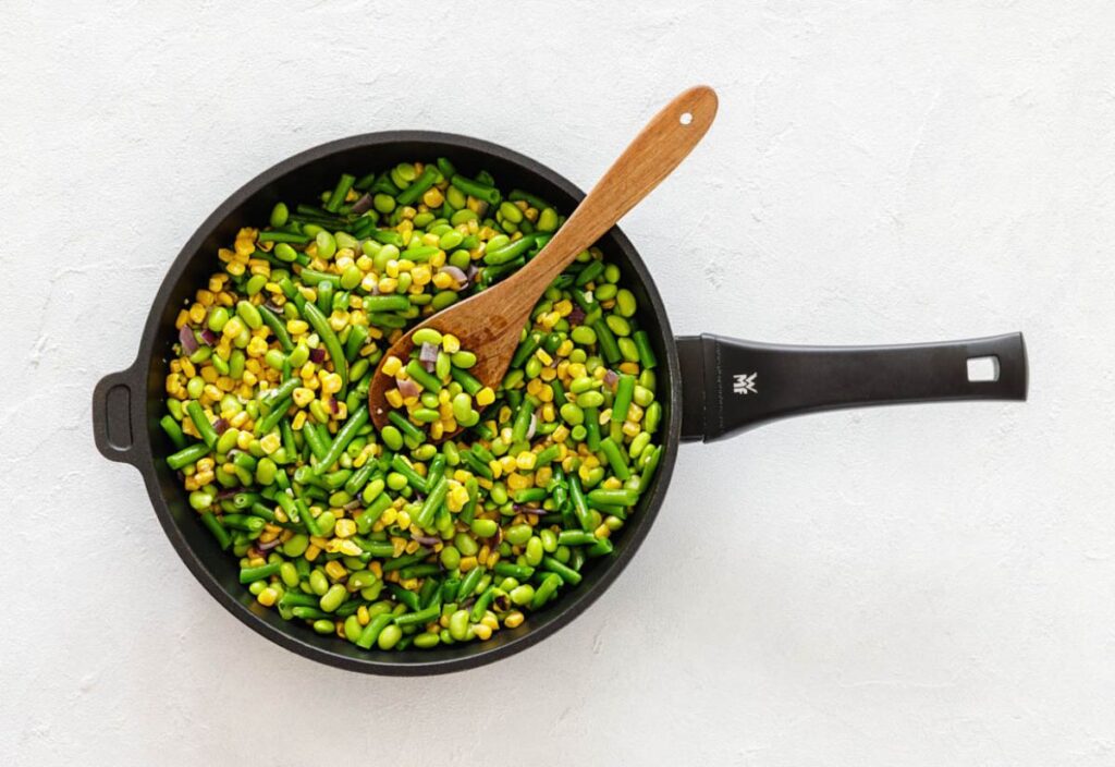 A skillet full of lima beans, corn, green beans, and visible onion pieces with a wooden spoon.