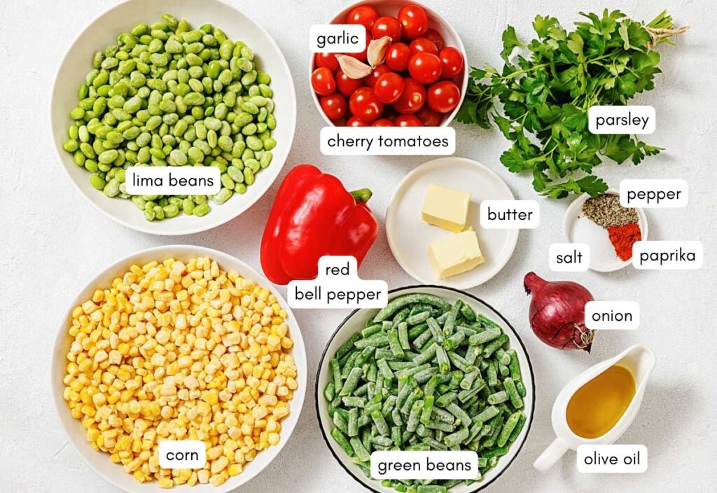 The top view of all the ingredients needed to make succotash includes lima beans, corn, green beans, cherry tomatoes, red bell pepper, garlic, onion, butter, parsley, salt, pepper, paprika, and olive oil.