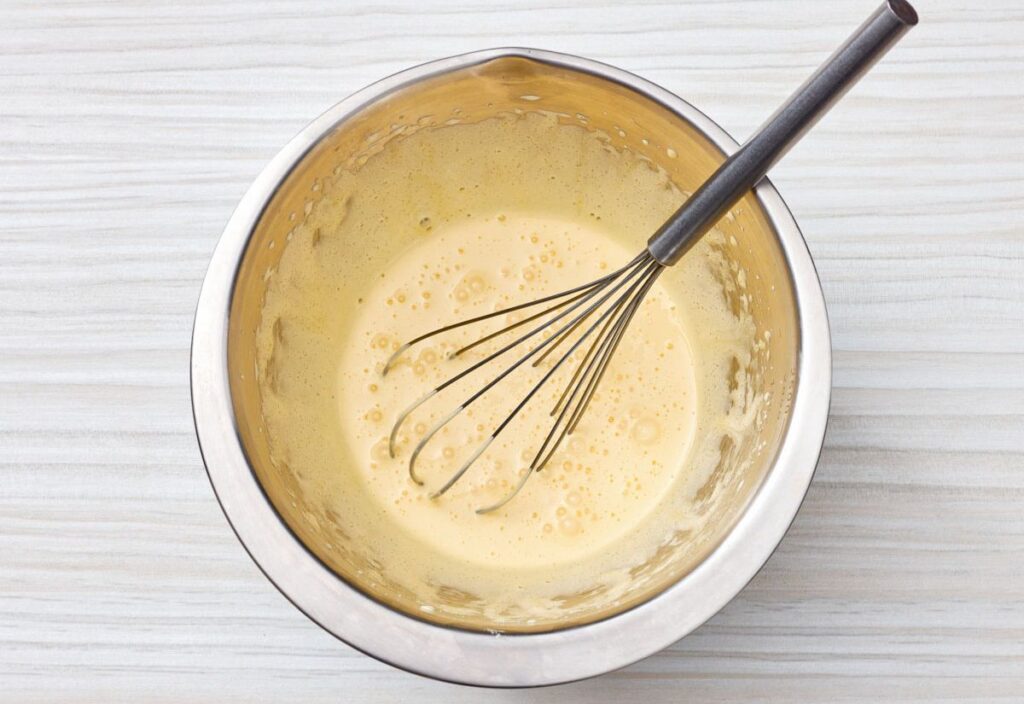 A metal mixing bowl containing whisked egg yolks, with bubbles forming on the surface, and a whisk resting inside the bowl.