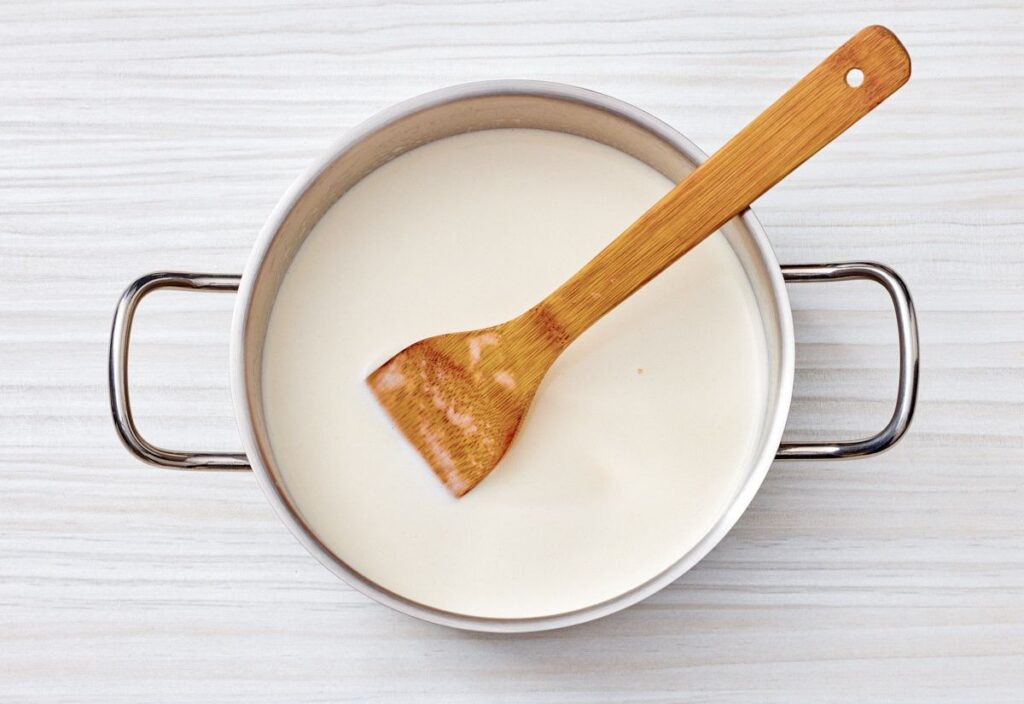 A bamboo spoon stirring milk in a saucepan on a light-colored wooden surface.