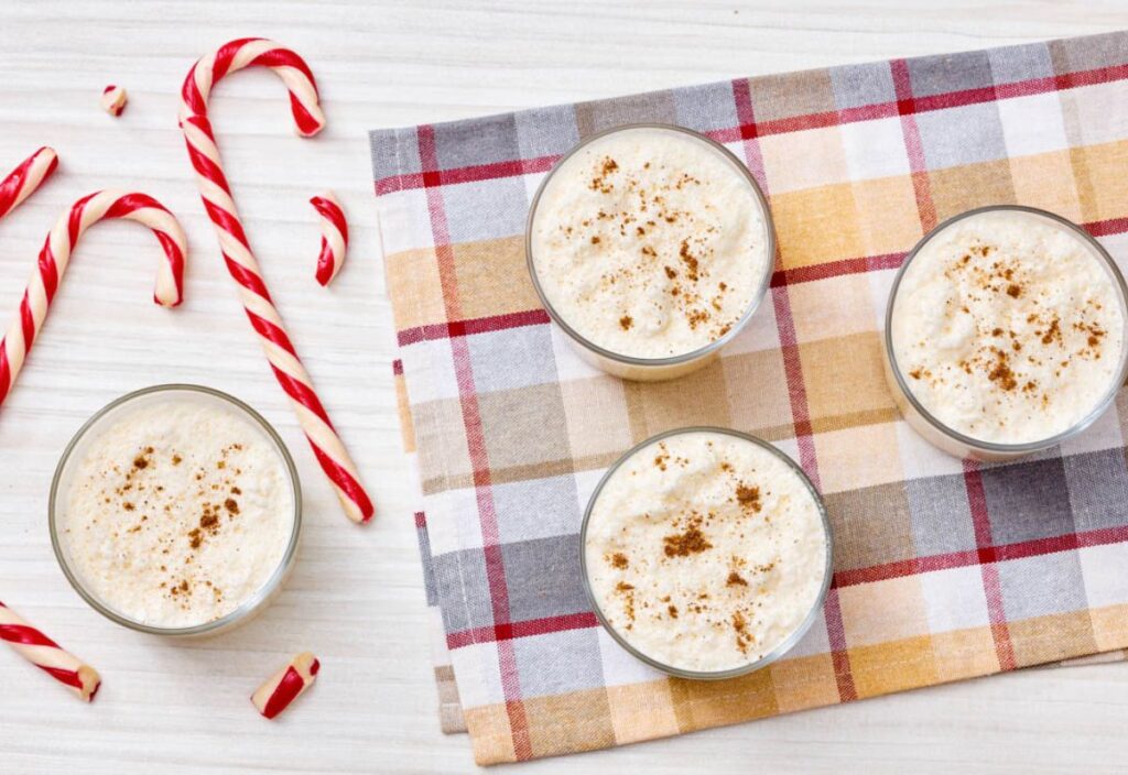 Four glasses of creamy eggnog topped with cinnamon, surrounded by red-and-white candy canes on a festive checkered cloth.