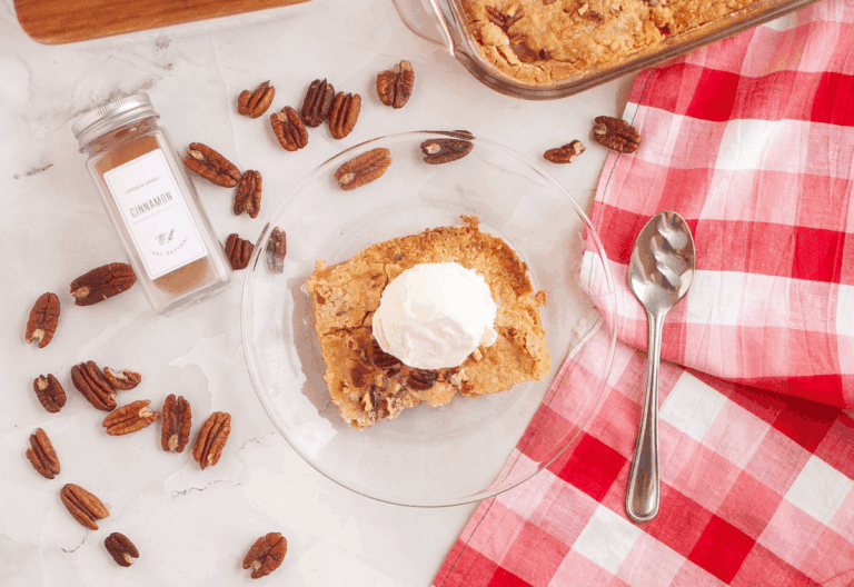 A serving of apple dump cake with a scoop of whipped cream on top, surrounded by pecans and a cinnamon jar, placed on a red and white checkered napkin.