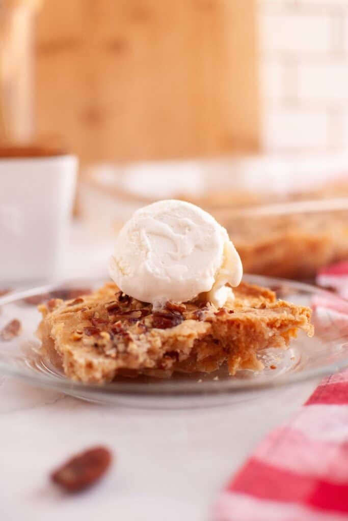 A serving of apple dump cake with a scoop of whipped cream on top, surrounded by pecans and a cinnamon jar, placed on a red and white checkered napkin.
