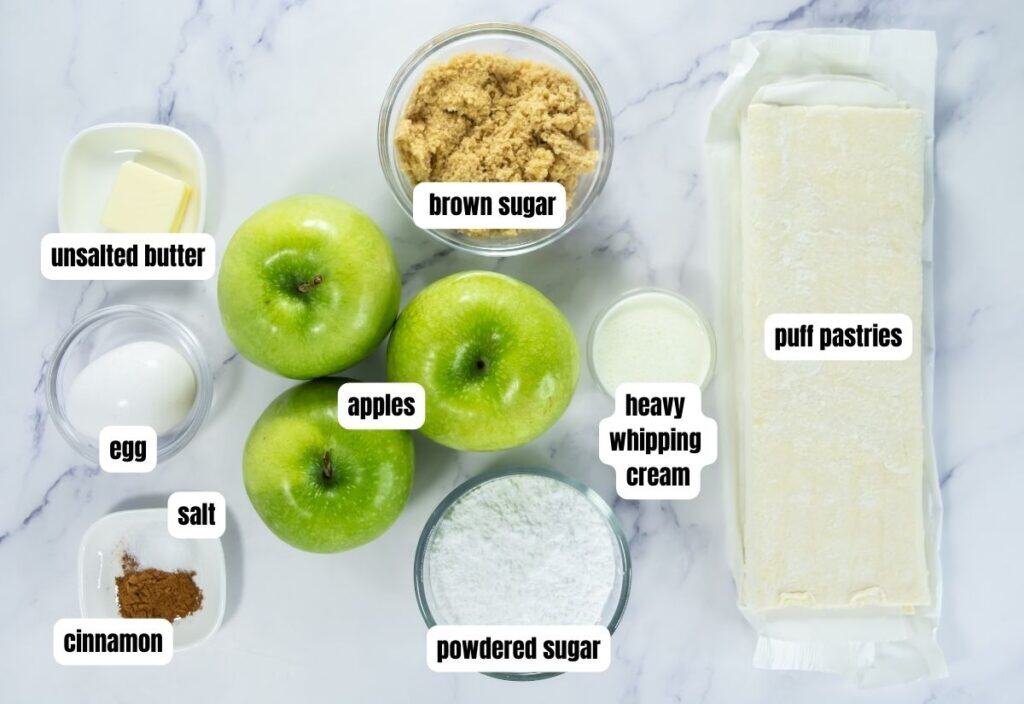 Top view of apple turnover ingredients neatly arranged on a white surface: puff pastry sheets, green apples, brown sugar, powdered sugar, heavy whipping cream, unsalted butter, egg, cinnamon, and salt, each labeled.