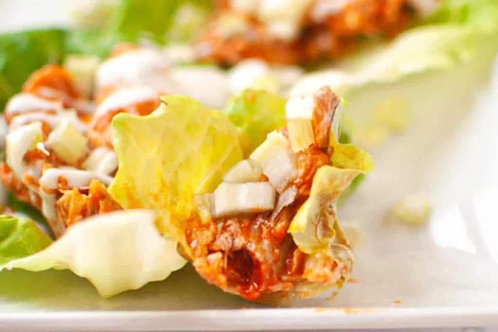 Close-up of a lettuce leaf topped with shredded meat in sauce, diced onions, and a drizzle of white dressing, on a white plate.