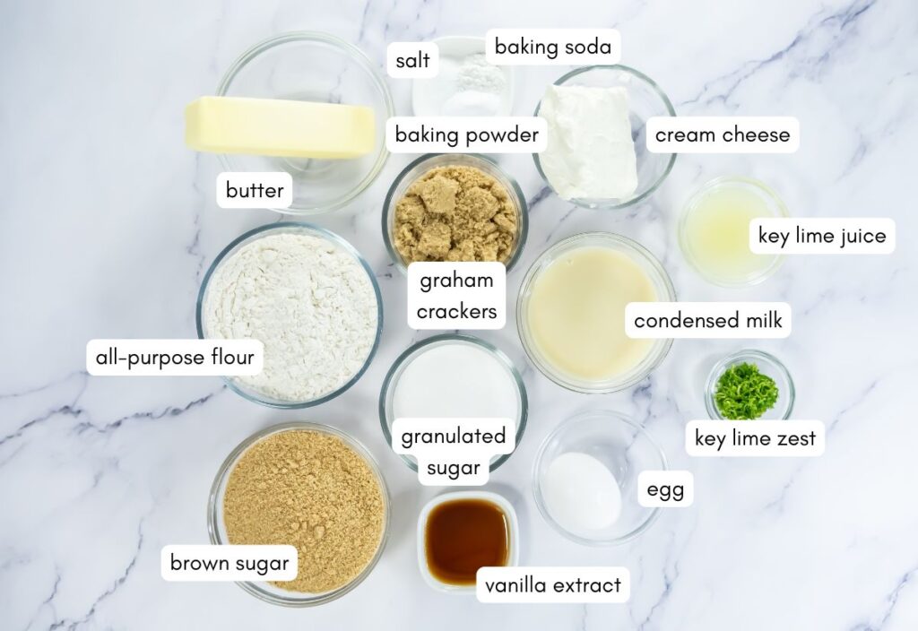 Labeled baking ingredients on a marble surface include butter, flour, brown sugar, vanilla, salt, baking powder, baking soda, cream cheese, lime juice, lime zest, condensed milk, milk, sugar, egg, and graham crackers.