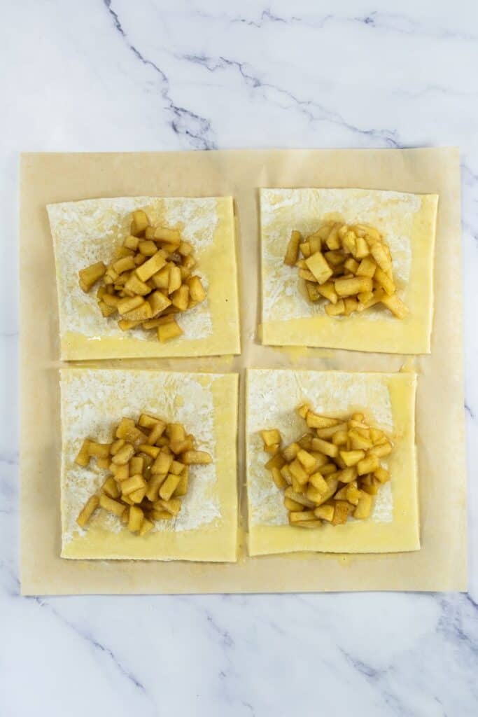 Puff pastry squares with cooked cinnamon apples, edges brushed with egg wash, and ready to be folded into turnovers.