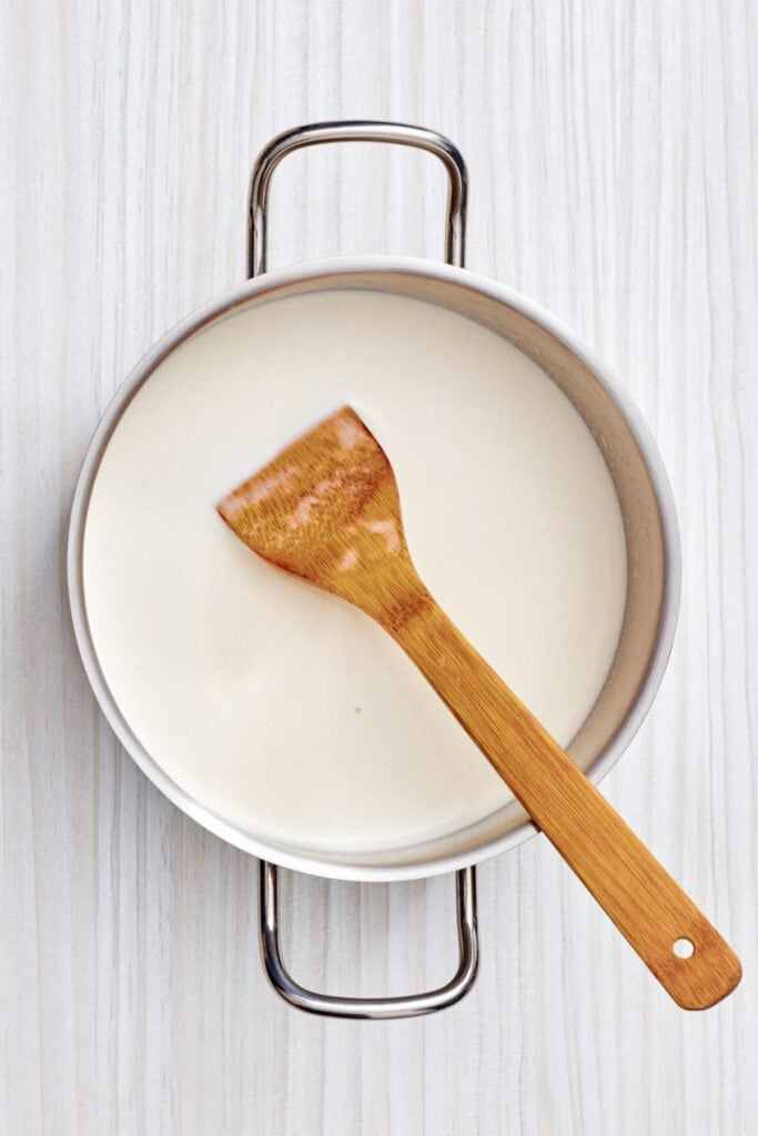 A bamboo spoon stirring milk in a saucepan on a light-colored wooden surface.