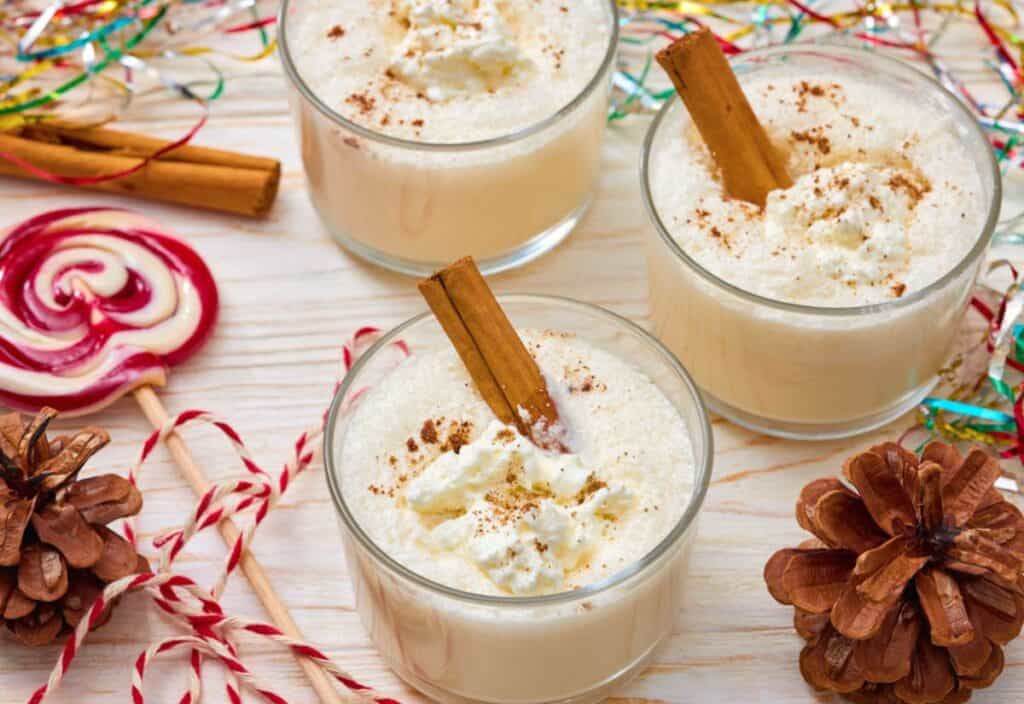 Three glasses of homemade egg nog topped with whipped cream and a cinnamon stick, surrounded by festive decorations like pinecones, candy canes, and colorful tinsel.