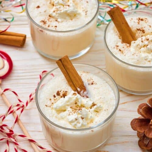 Three glasses of homemade egg nog topped with whipped cream and a cinnamon stick, surrounded by festive decorations like pinecones, candy canes, and colorful tinsel.