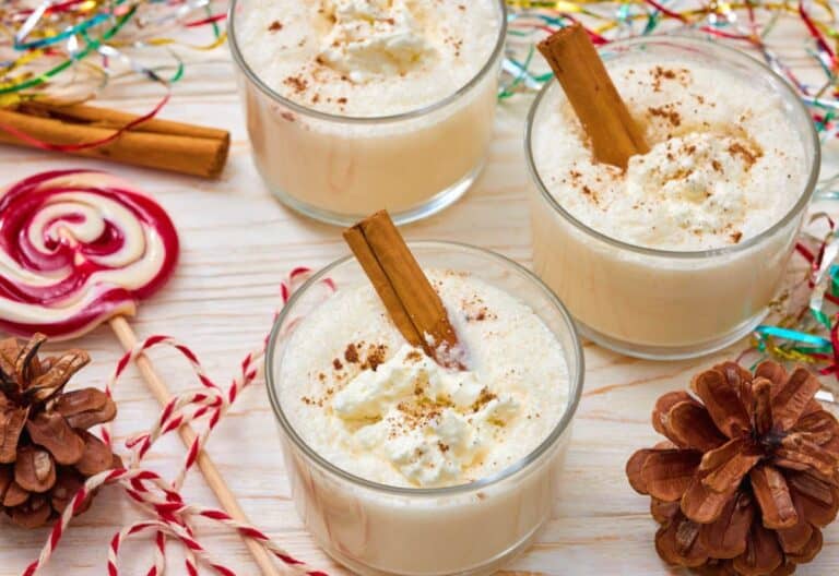 Three glasses of homemade egg nog topped with whipped cream and a cinnamon stick, surrounded by festive decorations like pinecones, candy canes, and colorful tinsel.