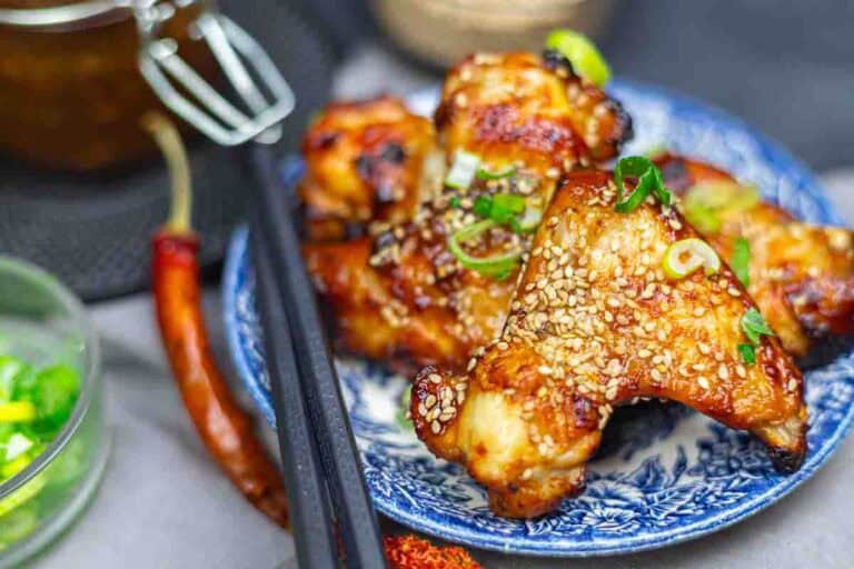 Grilled chicken wings glazed with sauce, topped with sesame seeds and green onions, served on a patterned blue plate with black chopsticks.