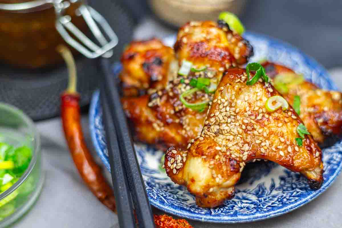 Grilled chicken wings glazed with sauce, topped with sesame seeds and green onions, served on a patterned blue plate with black chopsticks.