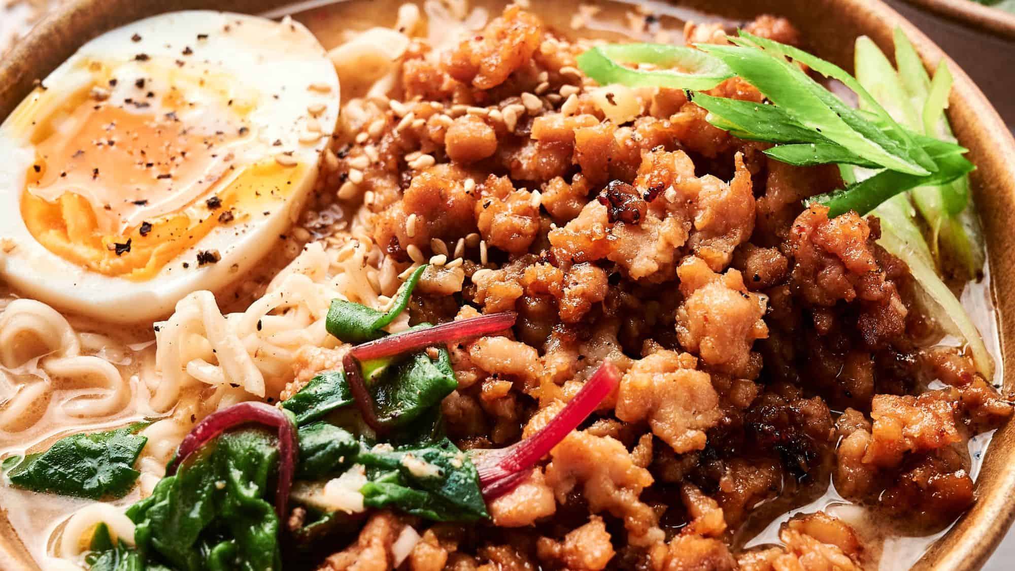 Close-up of a bowl of ramen with minced meat, spinach, sliced scallions, half a soft-boiled egg, and noodles in broth, garnished with sesame seeds and pepper.