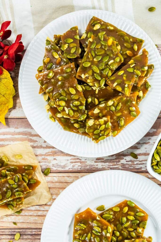 A plate of pumpkin seed brittle with a glossy caramel base, topped with green pepitas, surrounded by autumn leaves and a rustic wooden background.