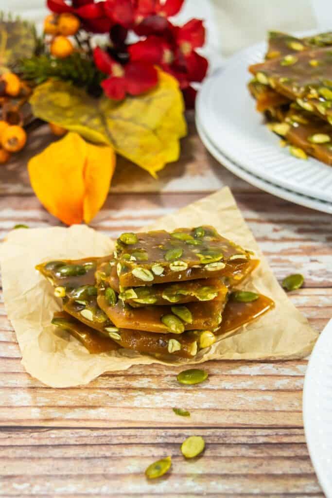 Close-up of stacked pumpkin seed brittle on parchment paper, with scattered pepitas, set against an autumn-themed backdrop of leaves and flowers.