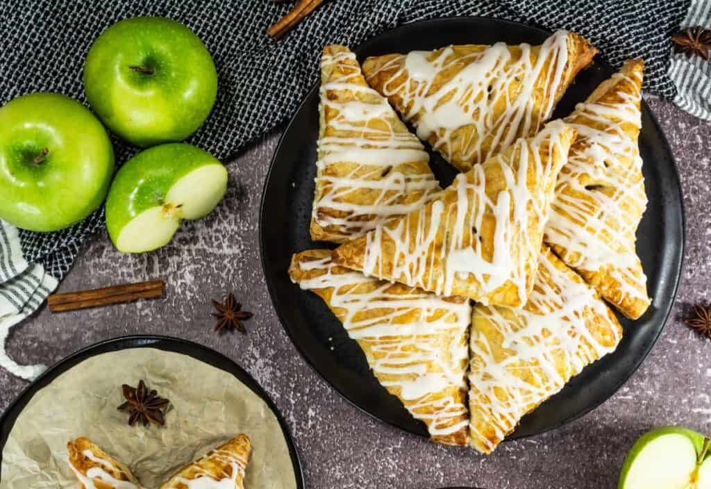Golden brown apple turnovers drizzled with white icing, stacked on a black plate, surrounded by fresh green apples, cinnamon sticks, and star anise on a textured surface.