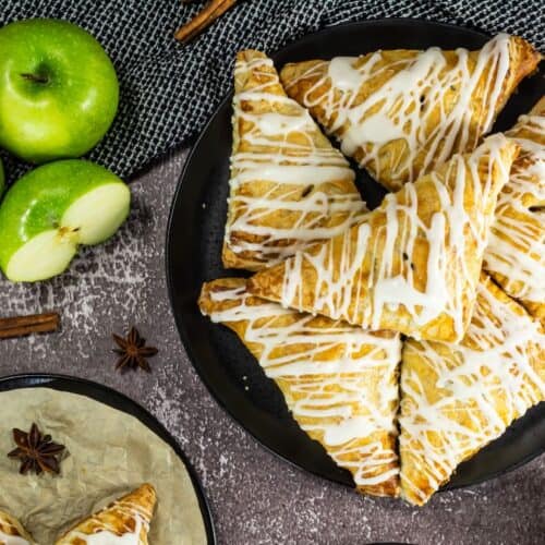 Golden brown apple turnovers drizzled with white icing, stacked on a black plate, surrounded by fresh green apples, cinnamon sticks, and star anise on a textured surface.