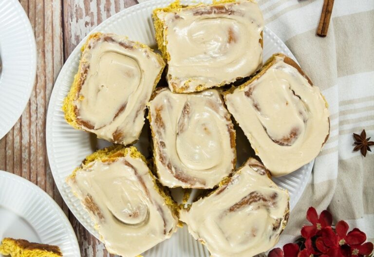 Seven golden-brown cinnamon rolls topped with creamy beige icing are arranged on a white plate. A striped cloth and rustic wood backdrop add warmth.