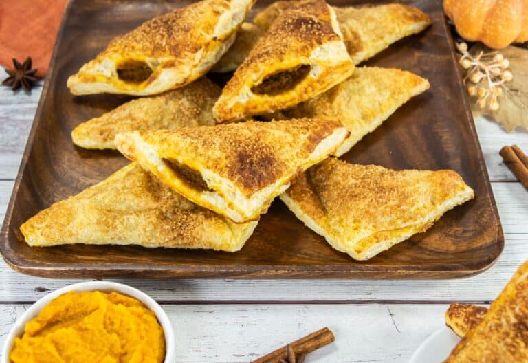Golden, flaky pumpkin turnovers arranged on a wooden plate with a bowl of pumpkin puree and cinnamon sticks nearby.