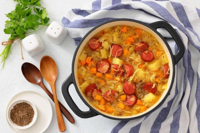 A pot of Chef Jenn’s Sauerkraut Soup with Kielbasa sits on a striped towel next to wooden spoons, bowls, fresh herbs, and salt and pepper shakers.