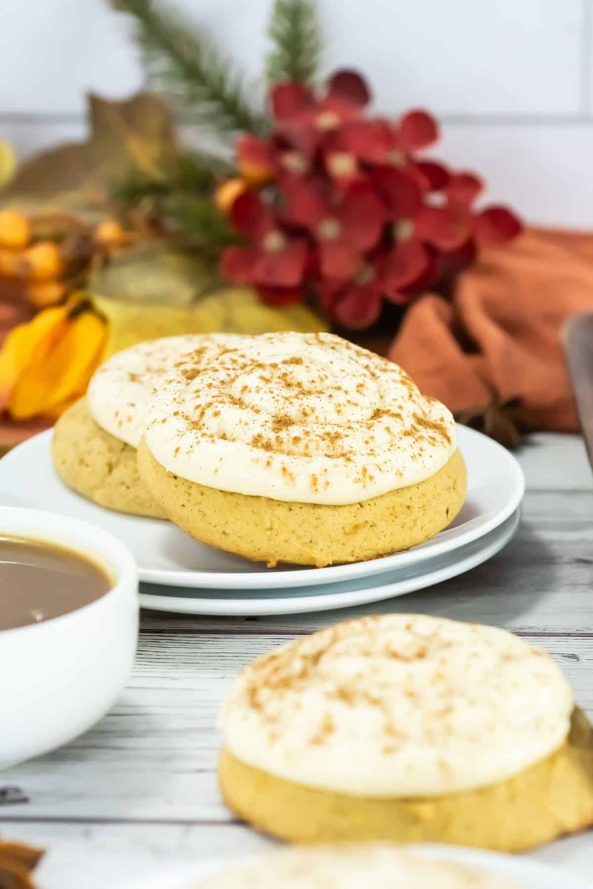 Frosted cookies with cinnamon on white plates, set on a wood table. A warm coffee cup is nearby, with autumn-themed leaves and flowers in the background.