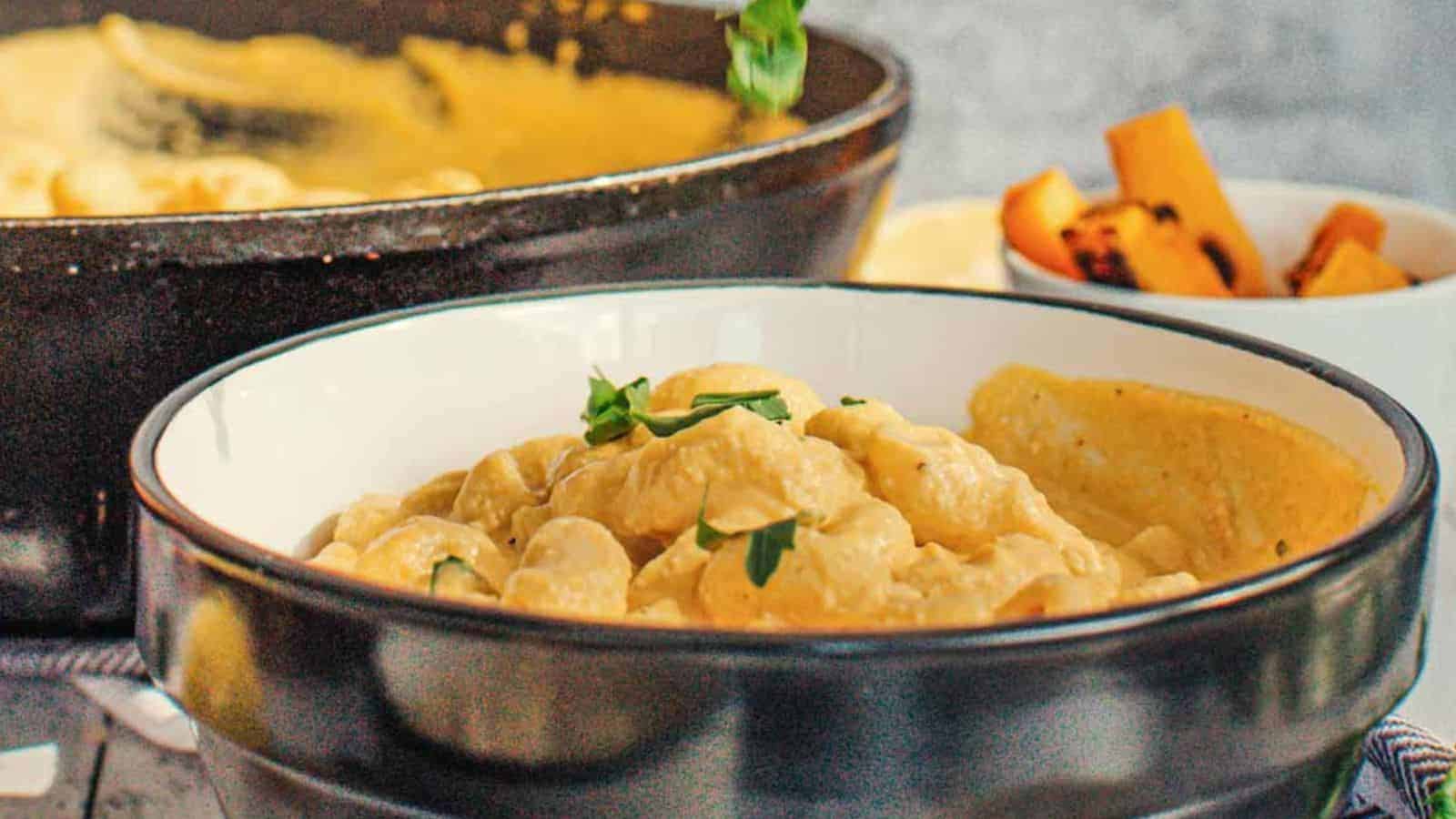 A bowl of creamy pasta garnished with herbs, with a slice of bread on the side; a skillet and a small bowl of roasted vegetables are in the background.