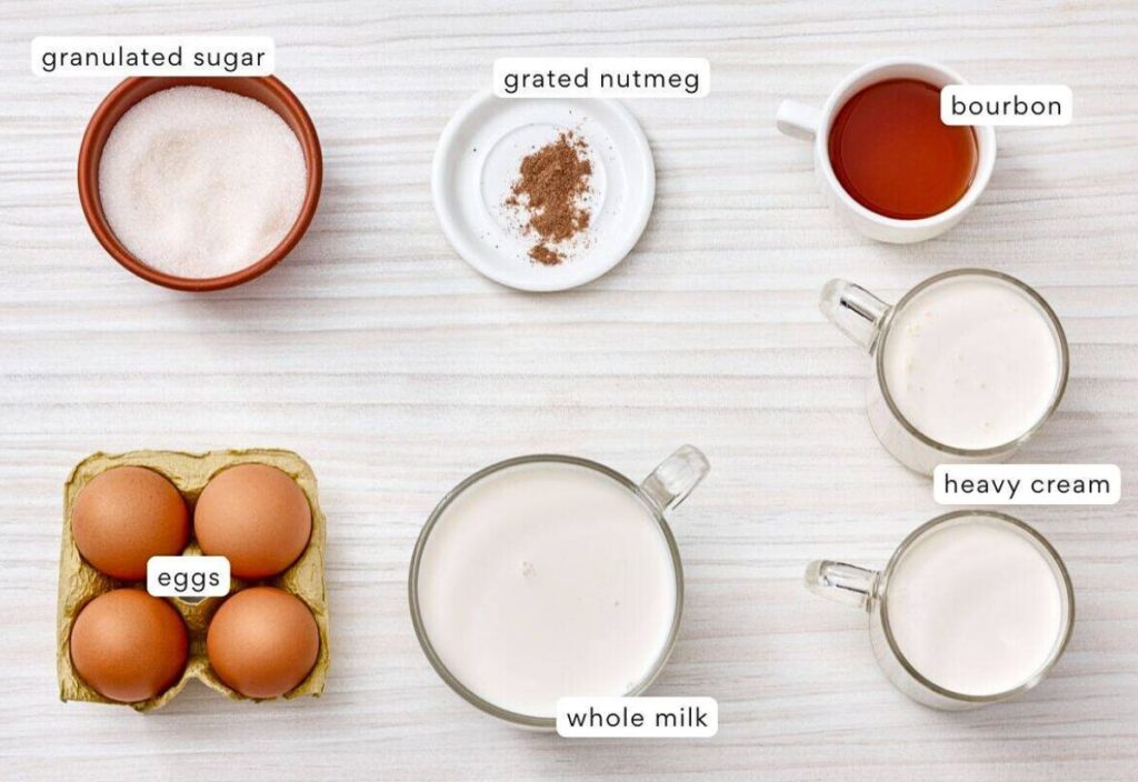 Ingredients for homemade egg nog: granulated sugar, grated nutmeg, liquor, large eggs, whole milk, and heavy cream arranged on a light wooden surface.