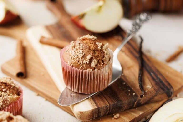 A muffin with crumb topping sits on a wooden board with a butter knife, surrounded by cinnamon sticks and apple slices.