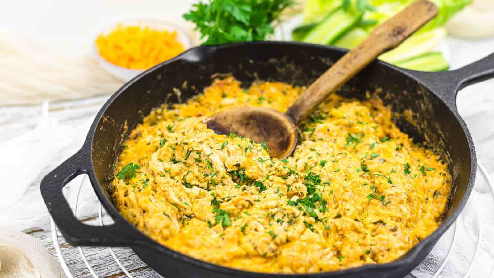 A cast iron skillet filled with a creamy dish, garnished with chopped herbs and a wooden spoon inside. Fresh parsley and sliced vegetables are seen in the background.