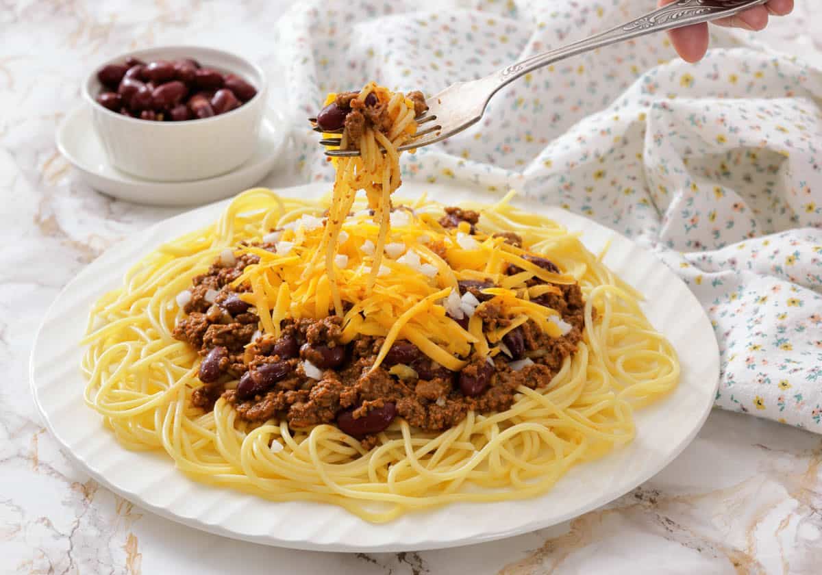 A plate of spaghetti topped with Cincinnati chili, shredded cheddar cheese, and chopped onions, with a fork lifting a bite.