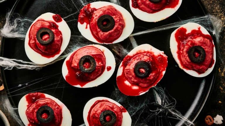 Deviled eggs topped with a black olive and red sauce, resembling bloody eyeballs, with spider webs around them.