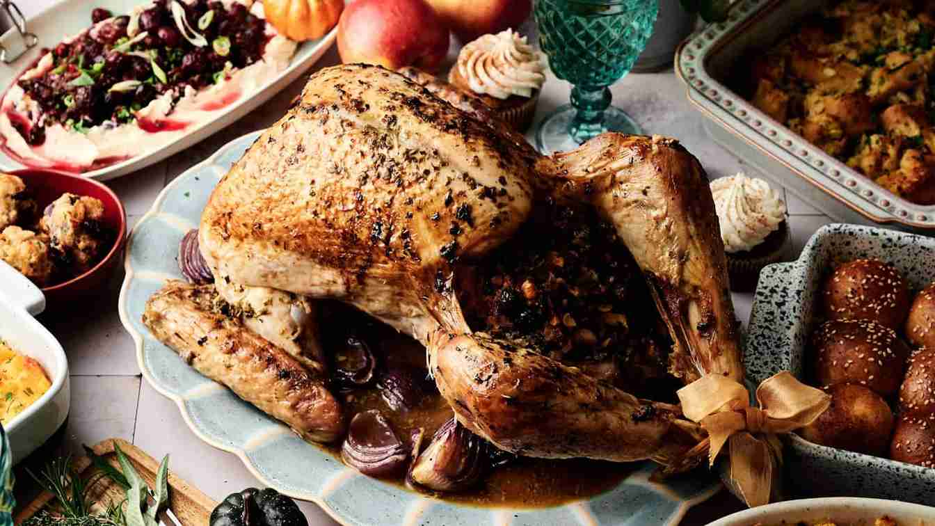 A roasted turkey stuffed with vegetables is served on a platter, surrounded by various side dishes including rolls, salad, and stuffing.