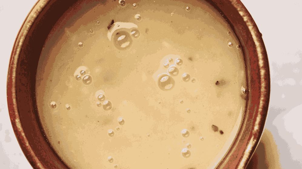 Close-up of a bowl filled with creamy turkey gravy soup, featuring small bubbles and herbs on the surface. The bowl has a reddish-brown rim.