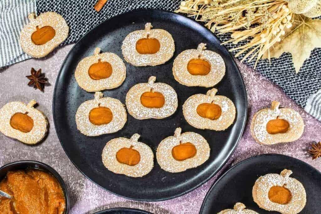 A black plate holds Pumpkin Linzer Cookies with powdered sugar and pumpkin filling. Other cookies and a bowl of filling are nearby on a textured tablecloth.