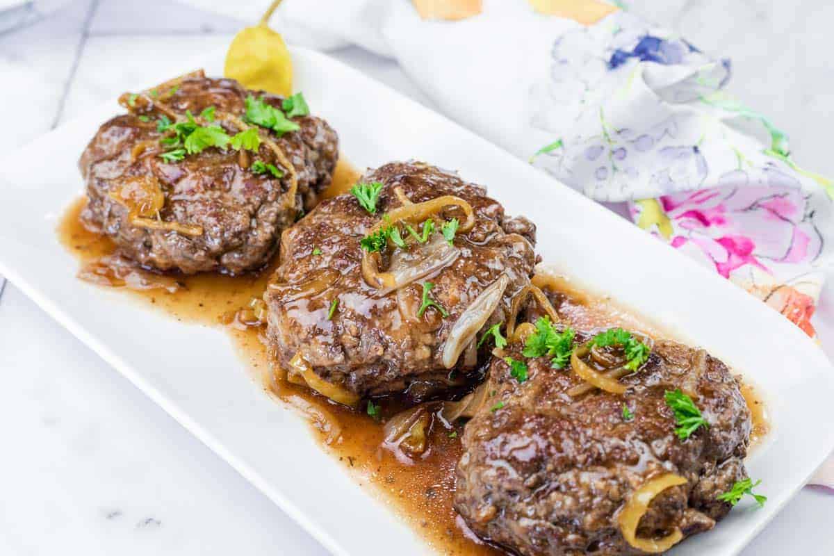 Three Salisbury steak patties topped with onions and brown gravy, garnished with parsley, served on a white rectangular plate.
