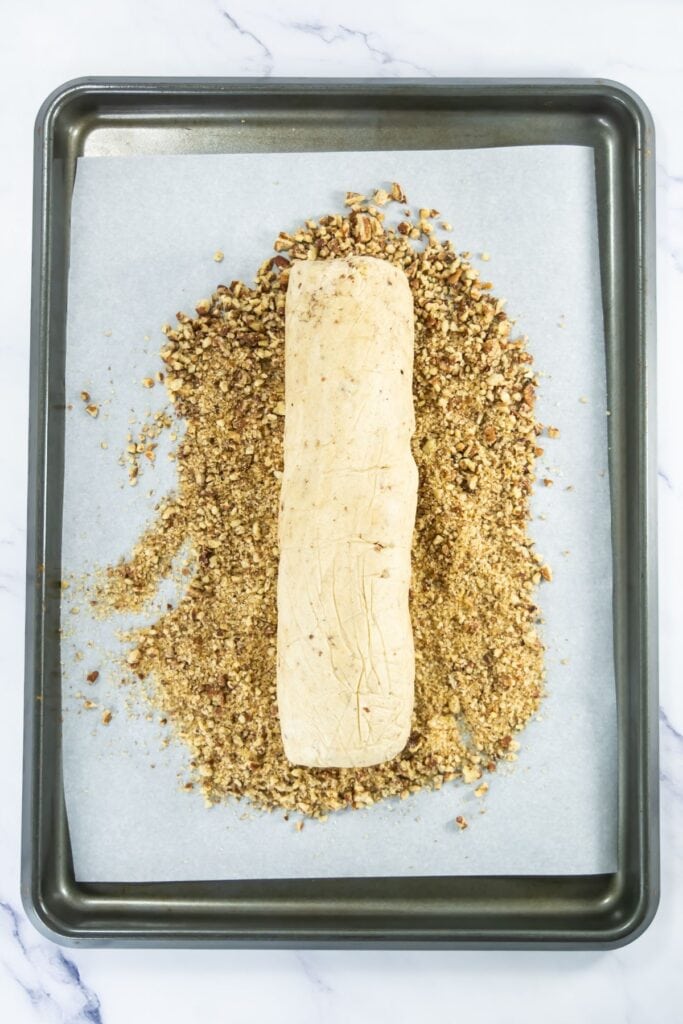 Cookie dough log on a parchment-lined baking sheet rolled in chopped pecans and sugar.
