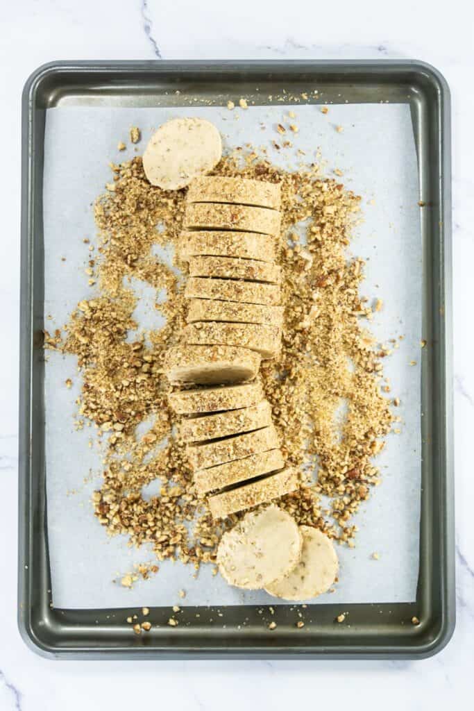 Coated pecan shortbread dough log sliced into rounds on a baking sheet, with crumbs scattered around.