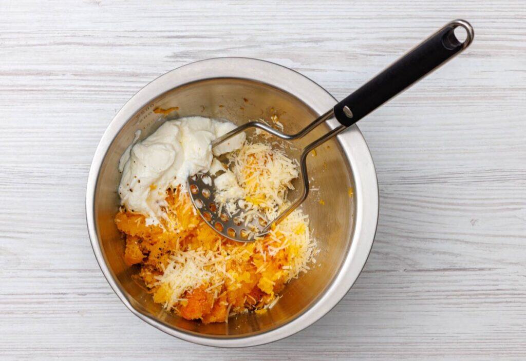 A bowl of cooked butternut squash mixed with Greek yogurt and grated Parmesan, with a potato masher inside.