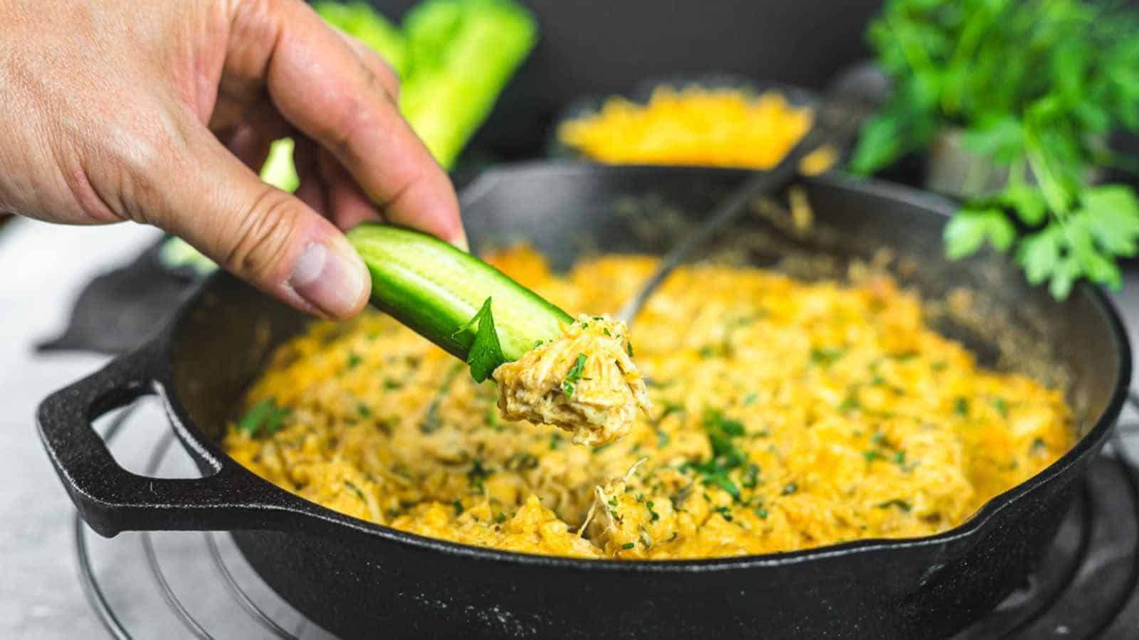 A cast iron skillet filled with a creamy dish, garnished with chopped herbs and a wooden spoon inside. Fresh parsley and sliced vegetables are seen in the background.
