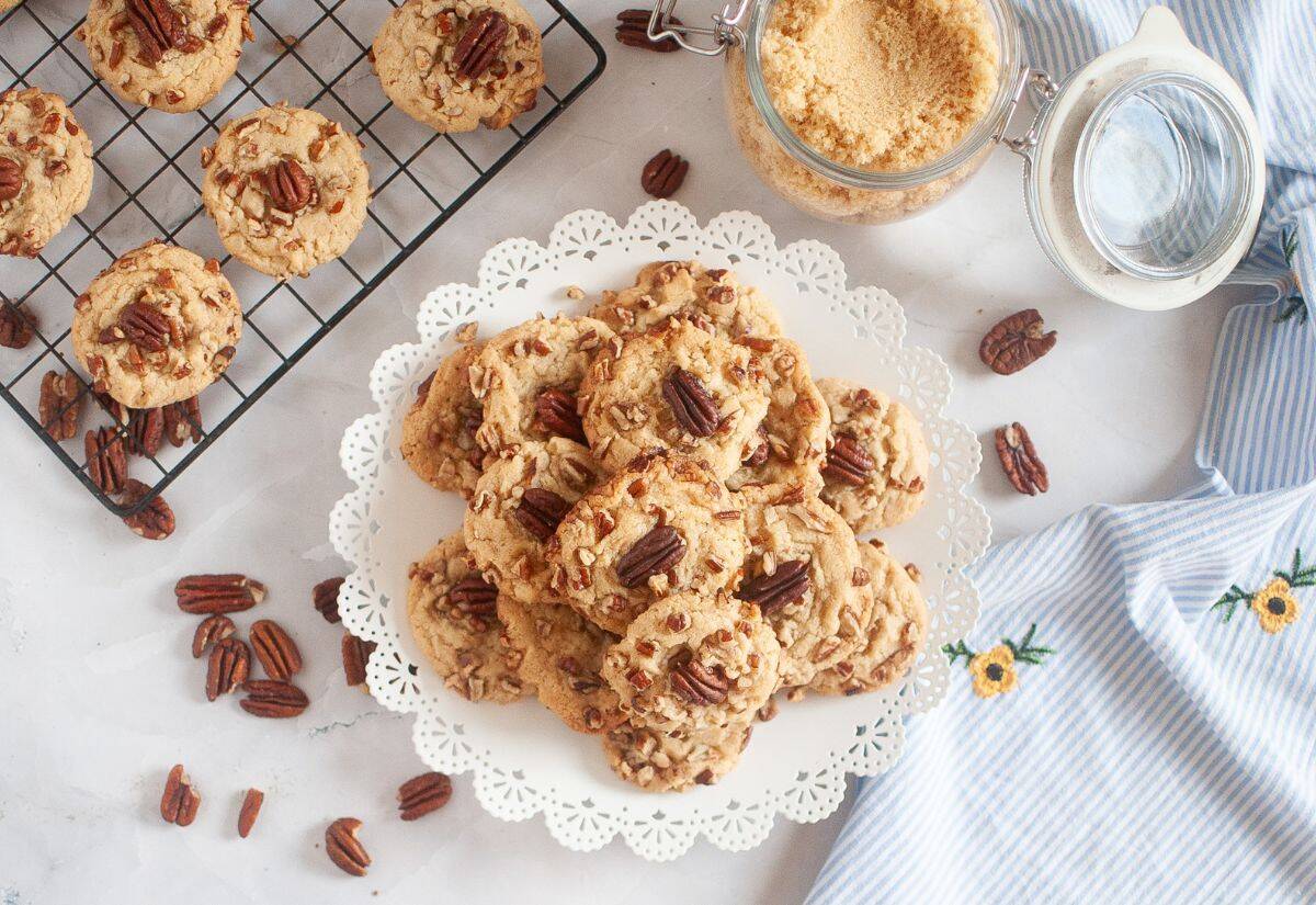 A plate of butter pecan cookies topped with pecan halves, surrounded by scattered pecans and cooling cookies on a rack.