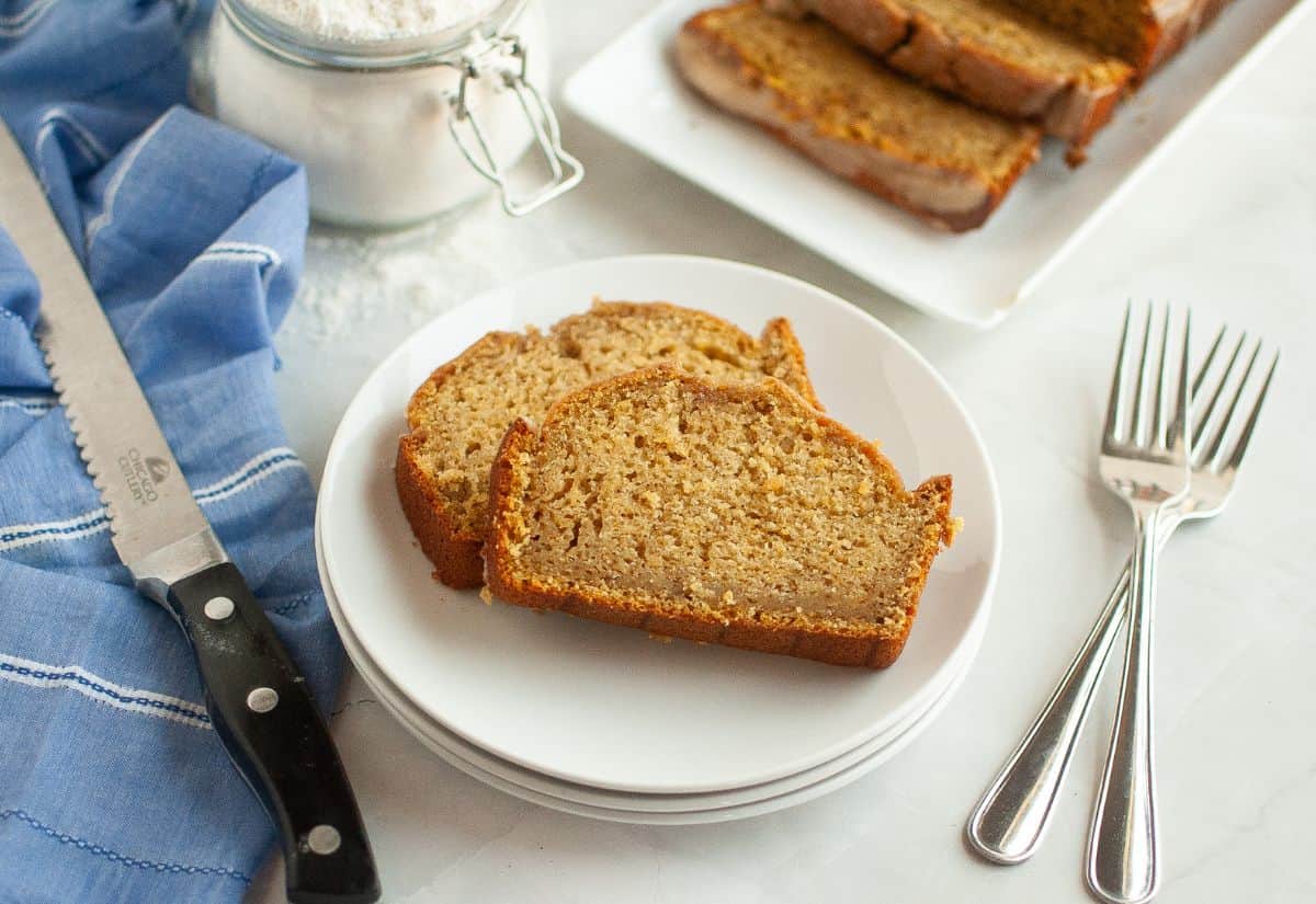 Two slices of butternut squash loaf on a white plate, with more slices on a tray in the background, a blue napkin, knife, and forks beside them.
