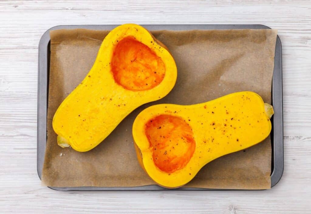 Two halved butternut squash pieces brushed with oil and seasoning on a parchment-lined baking sheet, ready for roasting.